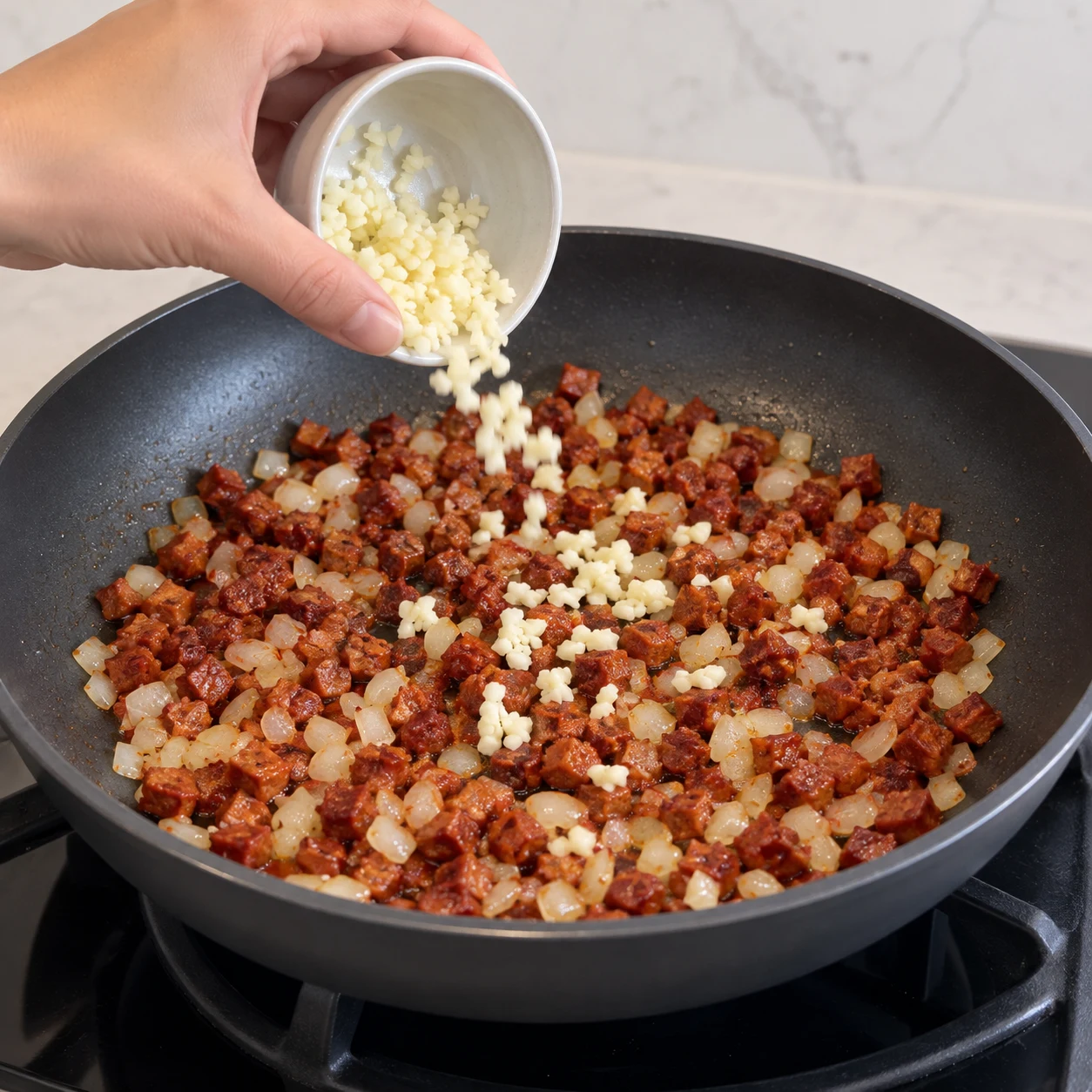 Une seule photo carrée montrant ajouter l’ail haché. L’ail est réparti et reste clair. Seulement les ingrédients déjà présents : Mini chorizo végétal, Mini chorizo végétal, Huile de tournesol, Oignon jaune, Ail. Les dés sont rouge-brun de chorizo entièrement végétal, sans viande ni morceaux de gras blanc.