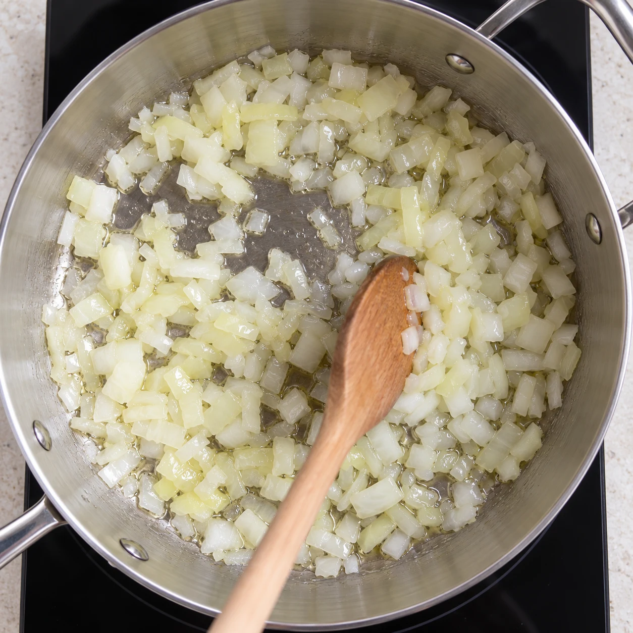 Oignon finement émincé revenant dans un filet d’huile d’olive dans une poêle inox sur une vraie plaque de cuisson; spatule en bois.