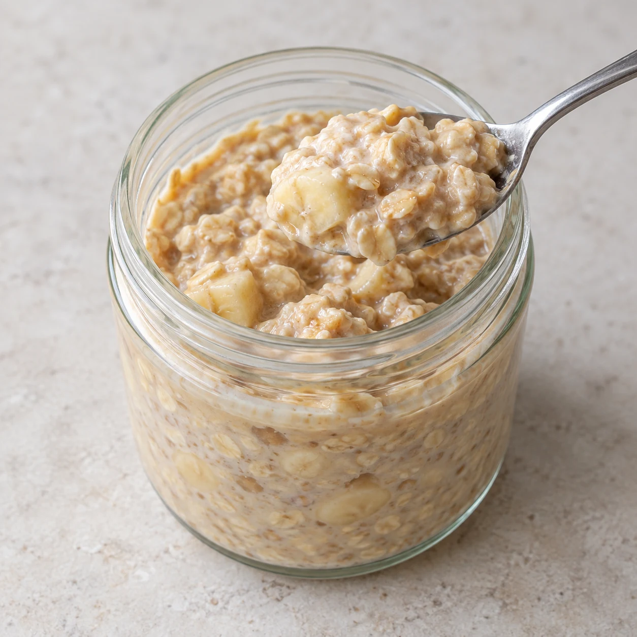 Un seul bocal ouvert après repos, avoine gonflée crémeuse avec morceaux de banane et teinte beige cacahuète ; une cuillère sans main.