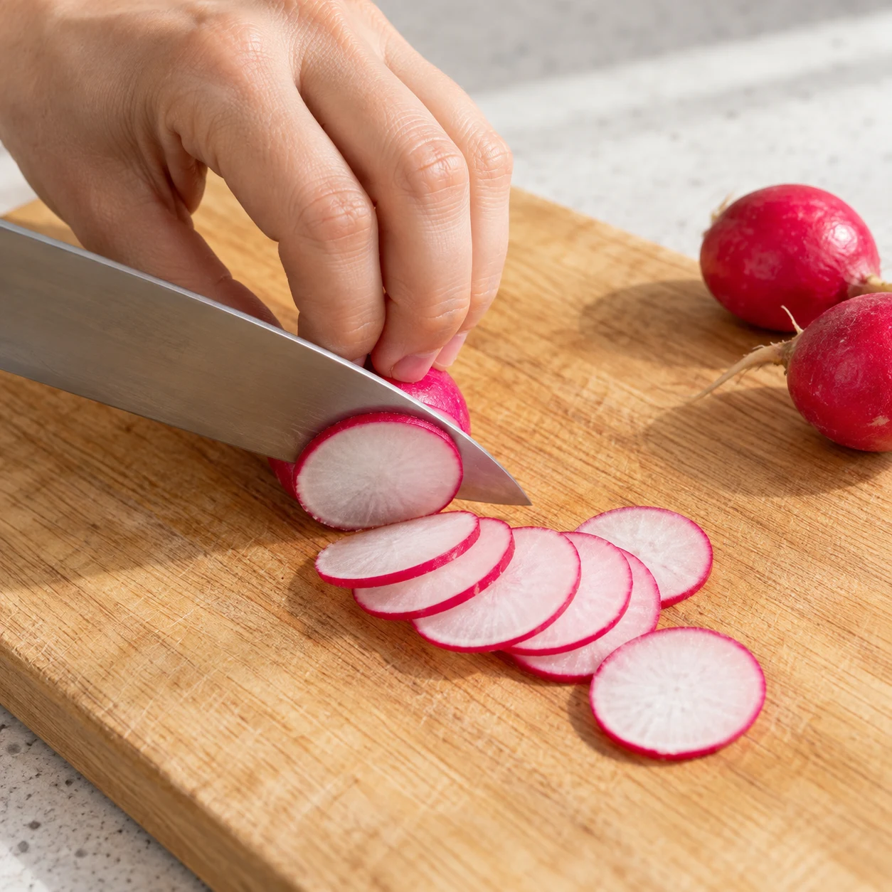 Couteau découpant un radis rose en fines rondelles sur une planche stable, main anonyme en griffe.