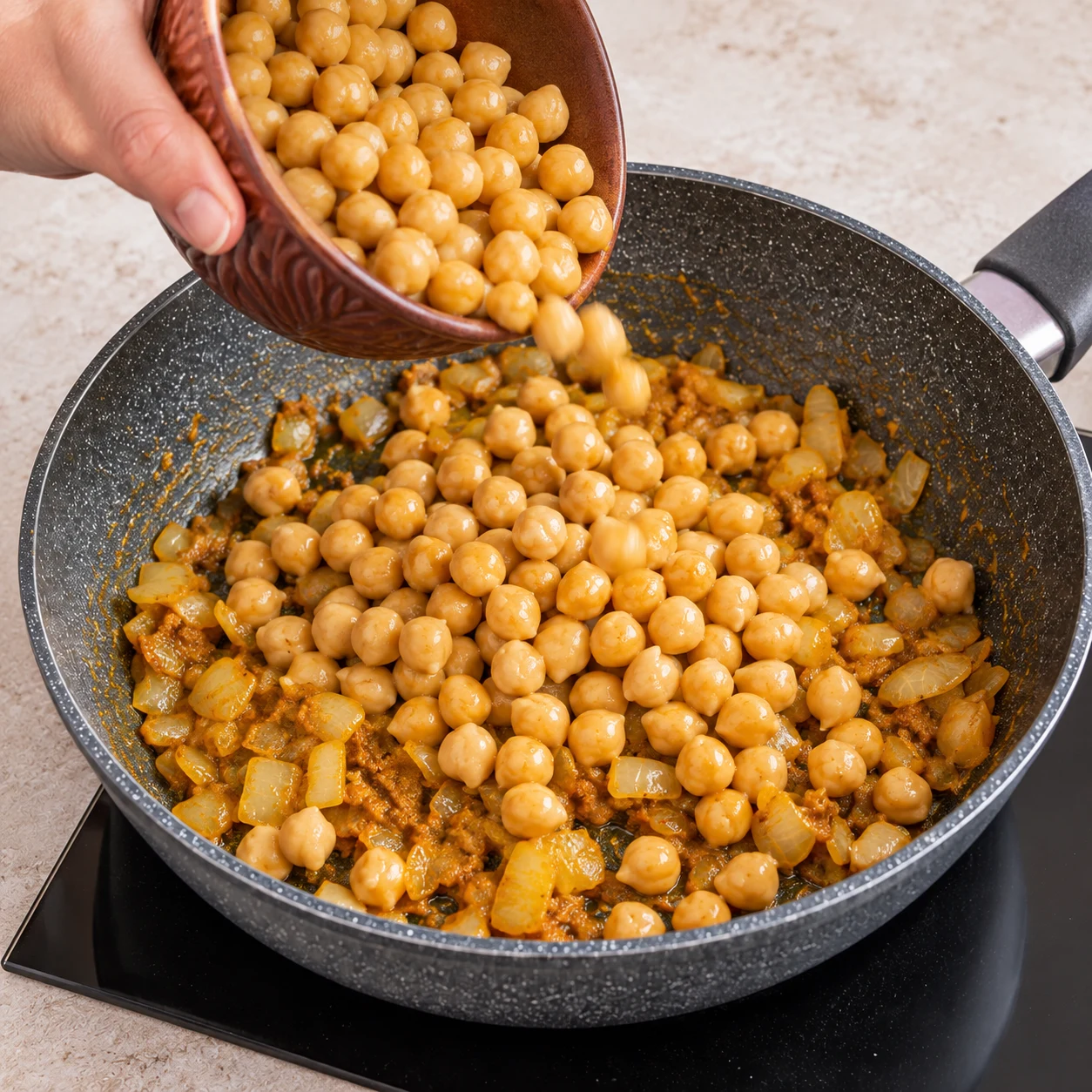 Main anonyme versant des pois chiches cuits dans la poêle avec oignon et pâte épicée.