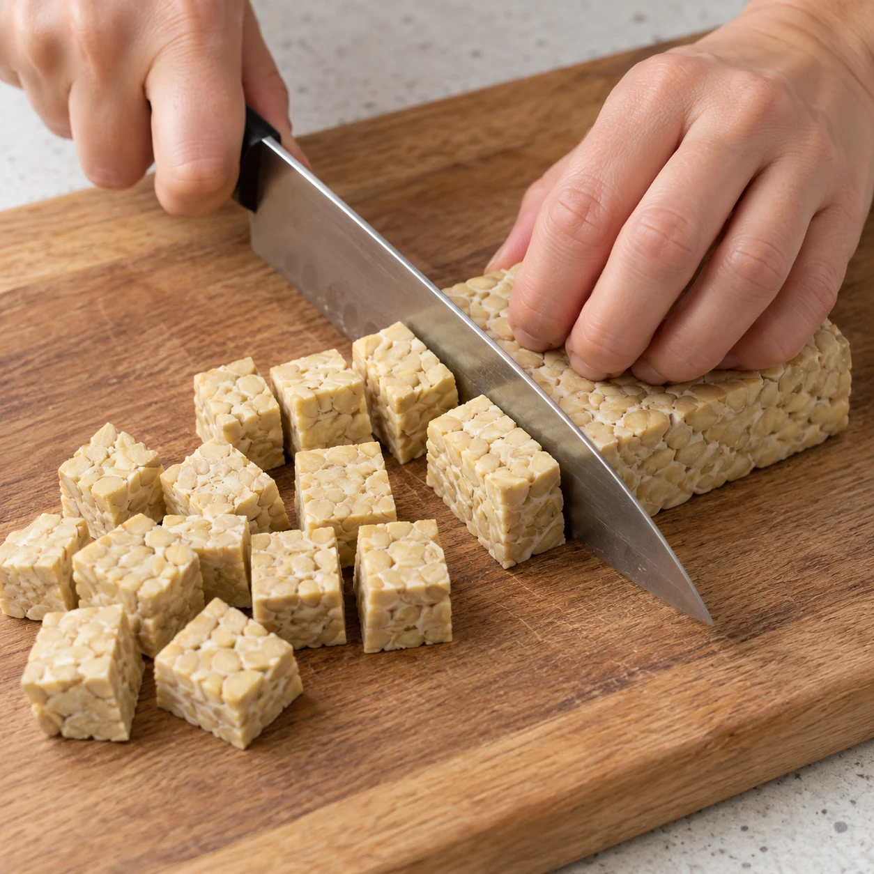 Mains adultes anonymes coupant un bloc de tempeh à grains de soja visibles en cubes; doigts en griffe.