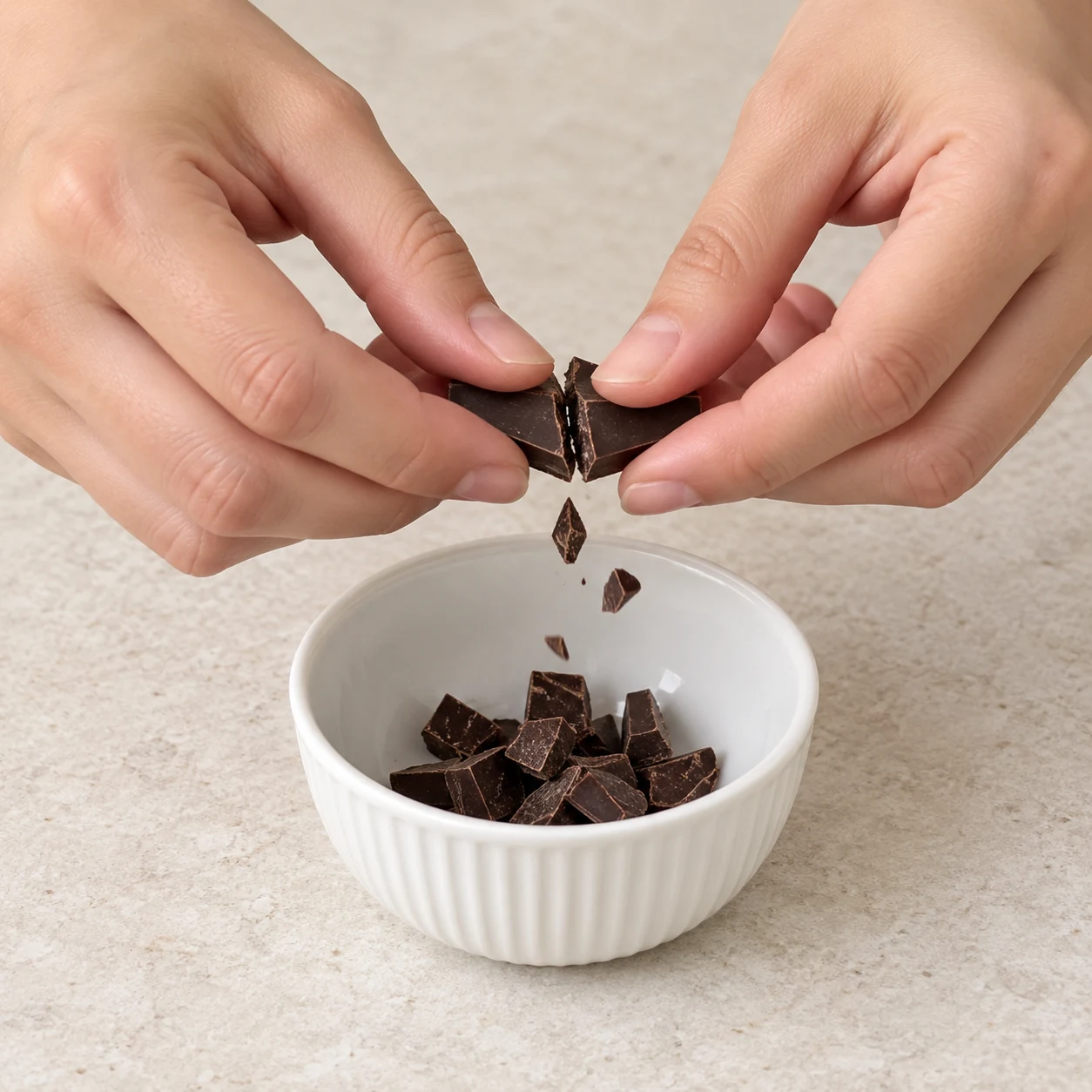 Mains adultes anonymes cassant un petit carré de chocolat noir au-dessus d’un bol.