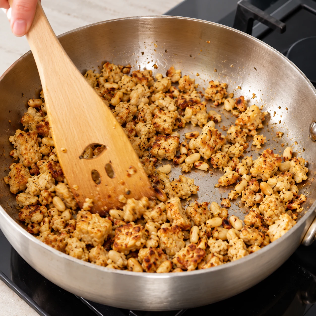 Spatule en bois remuant du tempeh émietté doré et des pignons dans une poêle en inox.