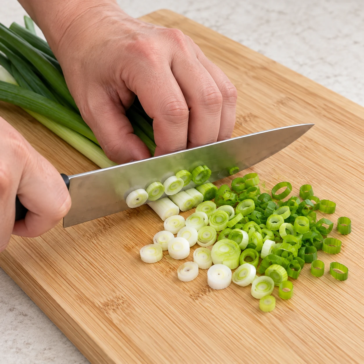 Coupez la branche de cébette en fines rondelles sur la planche, avec les doigts repliés. Résultat visuel : Le blanc et le vert de la cébette sont coupés en rondelles. Montrer seulement les ingrédients de cette étape, sur plan de travail pierre pâle ; pas de viande, pas d’œuf, pas de tofu.