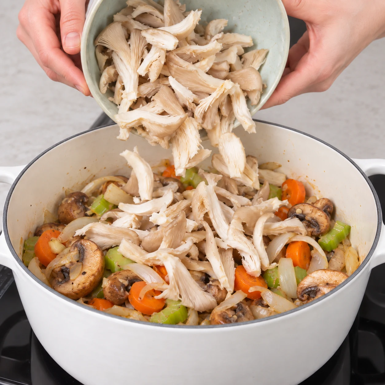 Des lanières fraîches de pleurotes beige clair tombent dans la cocotte contenant carotte, céleri, oignon et champignons bruns ; aucun jacquier.