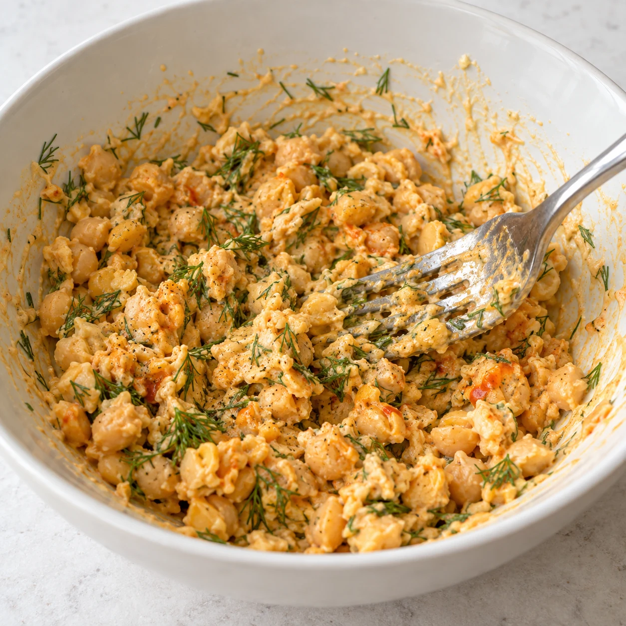 Fourchette mélangeant des morceaux de pois chiches écrasés enrobées d’une sauce légèrement orangée avec de fines frondes d’aneth, sans œufs ni jaune.
