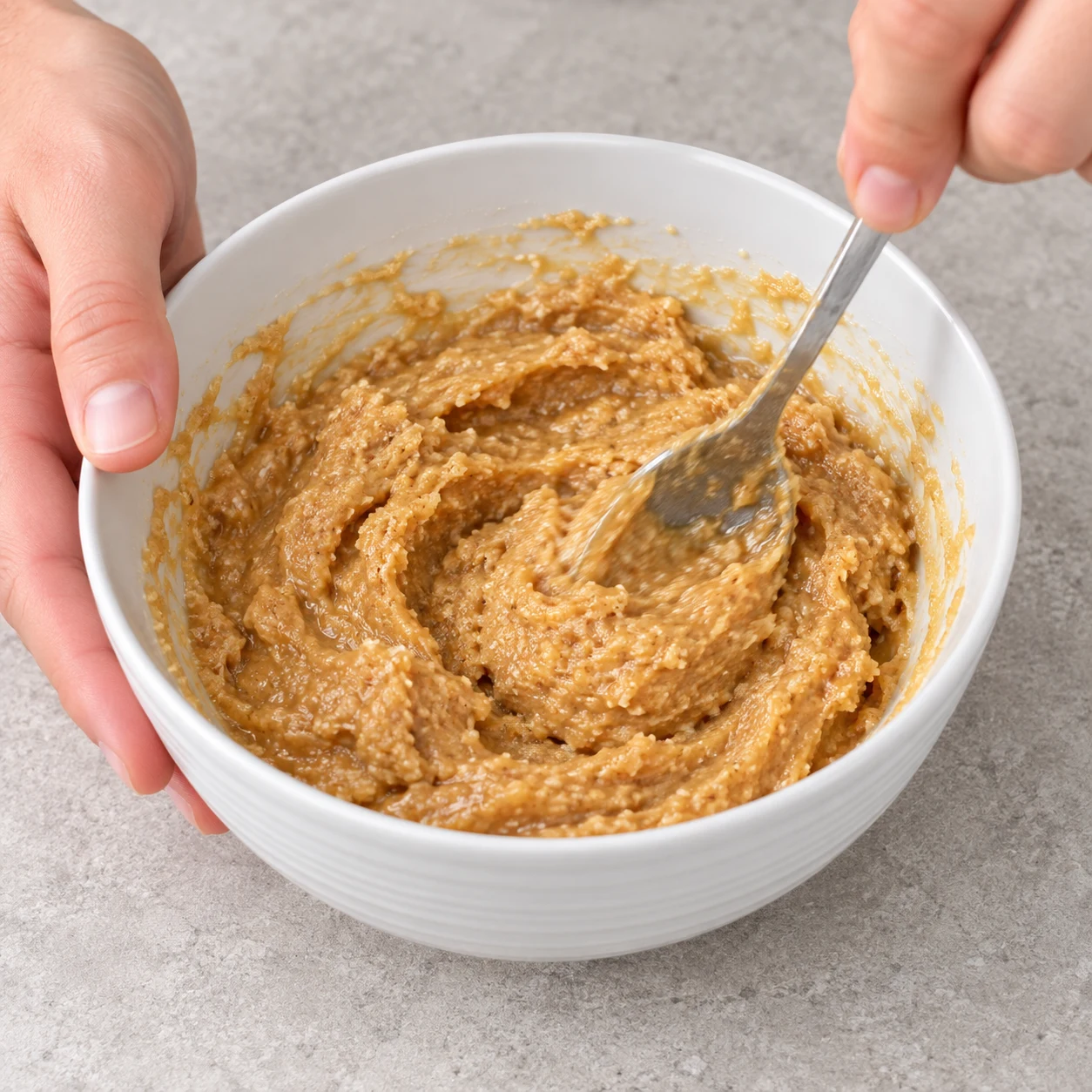 Une cuillère mélange la poudre d’amandes et le beurre de cacahuète avec le sirop dans un bol blanc.