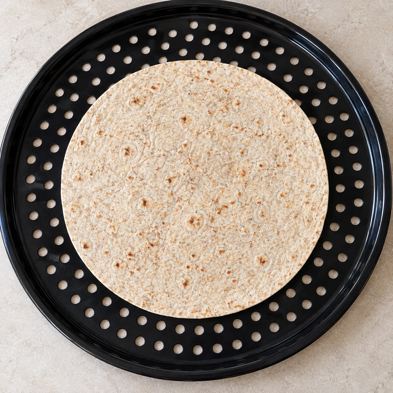 Une tortilla de blé crue et fine centrée sur une plaque ronde perforée noire.