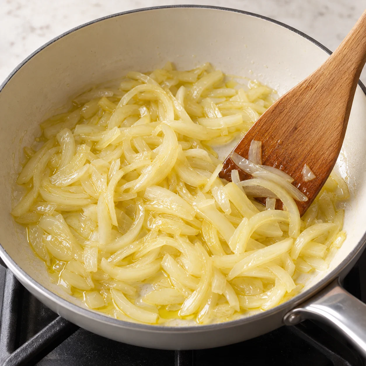 Photo culinaire carrée en lumière naturelle : oignon jaune émincé devenu translucide dans un filet d’huile d’olive au fond d’une poêle, aucun autre ingrédient visible, aucun texte, aucune marque.