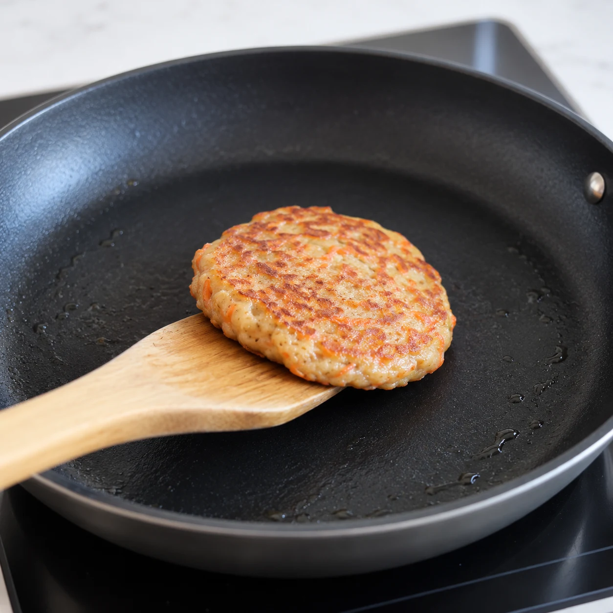 Spatule sous un petit pancake carotte au moment du retournement, première face dorée révélée. Poêle noire.