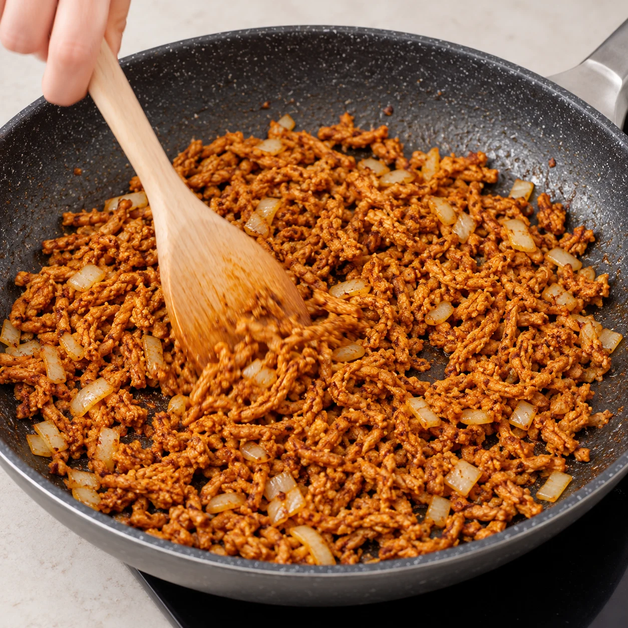 Haché végétal brun orangé remué à la cuillère en bois avec l’oignon doré dans une poêle noire.
