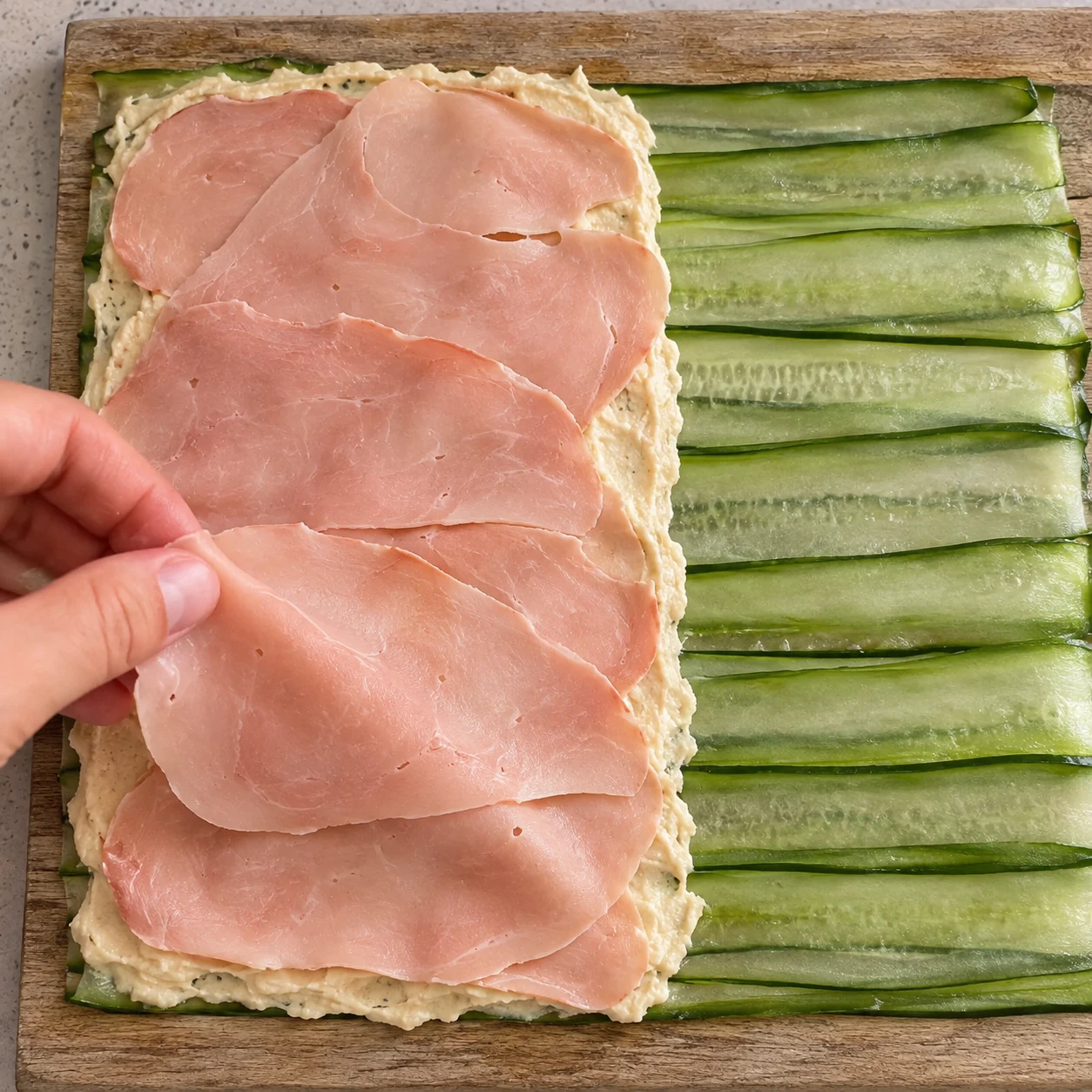 Déposer de fines tranches roses de jambon végétal sur le houmous beige couvrant la moitié de la nappe de concombre.