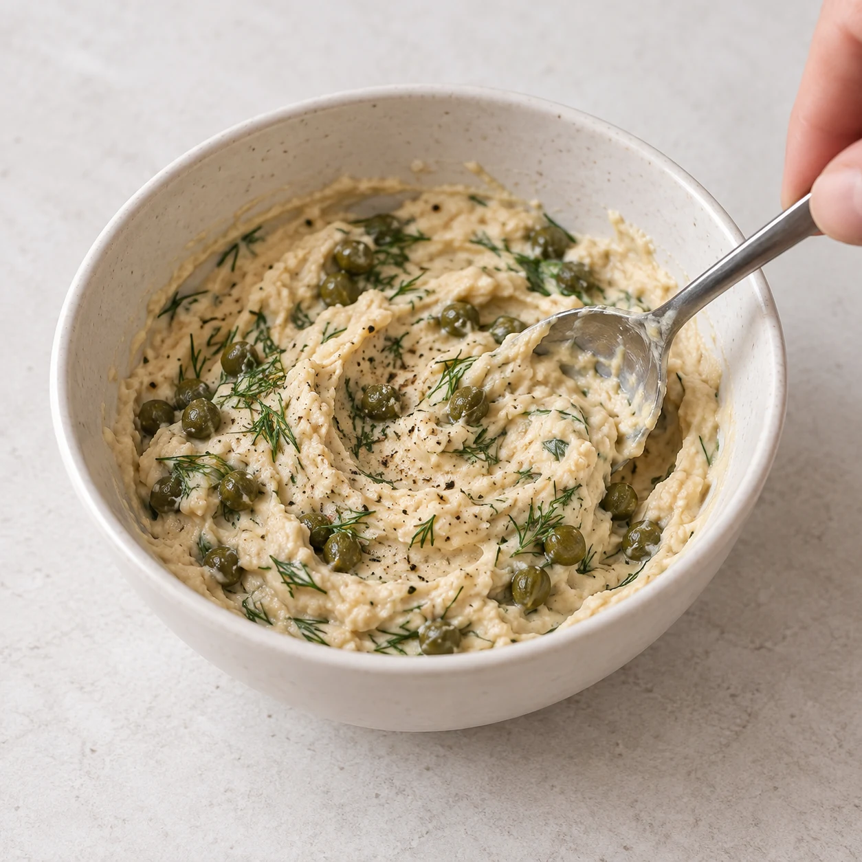 Houmous beige dense parsemé de petites câpres vertes et de brins d’aneth, remuée avec une cuillère.