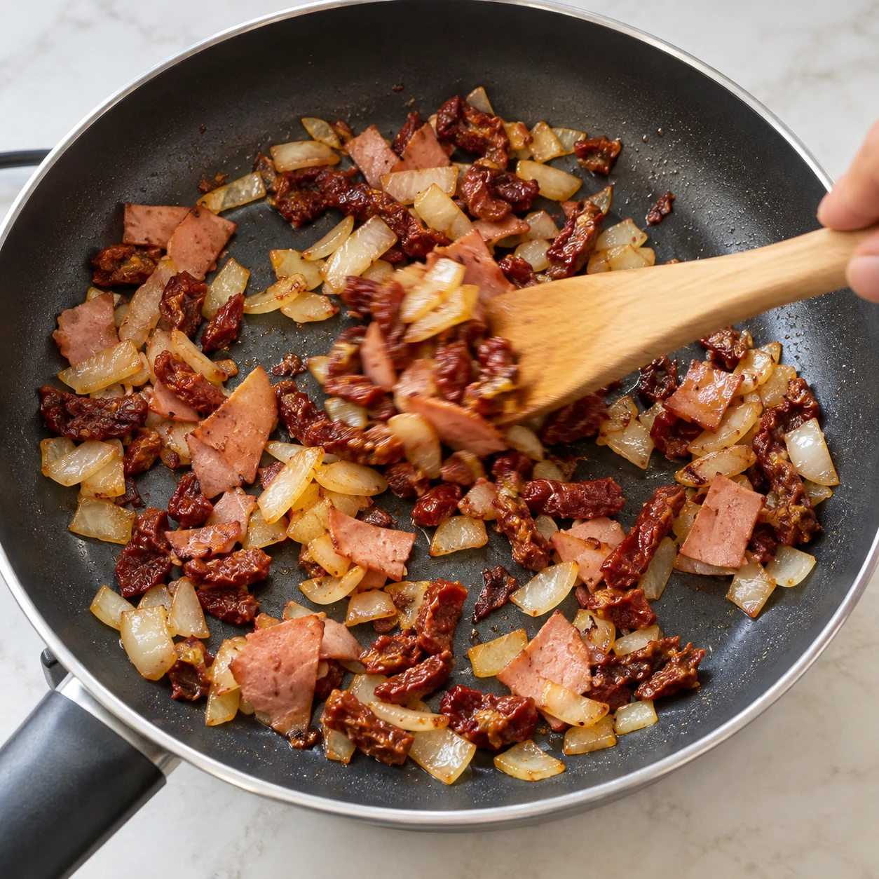 Spatule remuant ensemble oignon doré, morceaux de tomates séchées et jambon végétal dans une poêle antiadhésive.