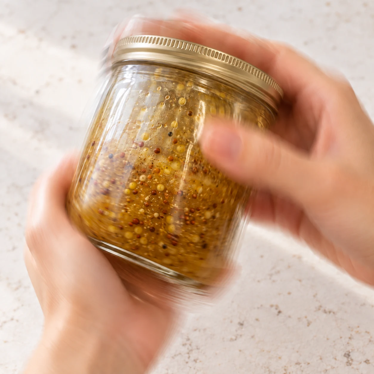 Deux mains anonymes secouent un bocal fermé, sauce homogène à la moutarde en grains.