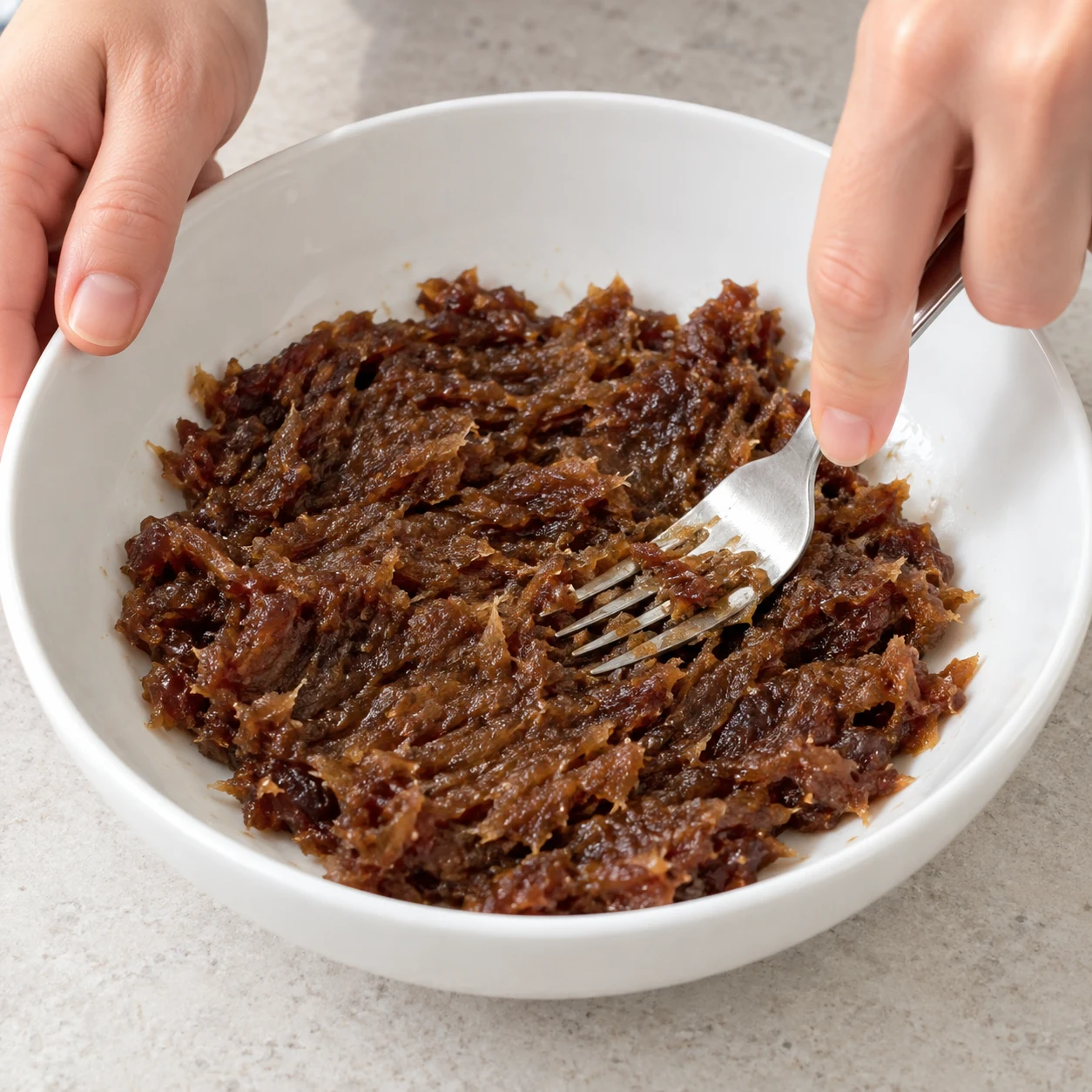 Fourchette écrasant les dattes en pâte brune fibreuse dans une assiette creuse blanche.