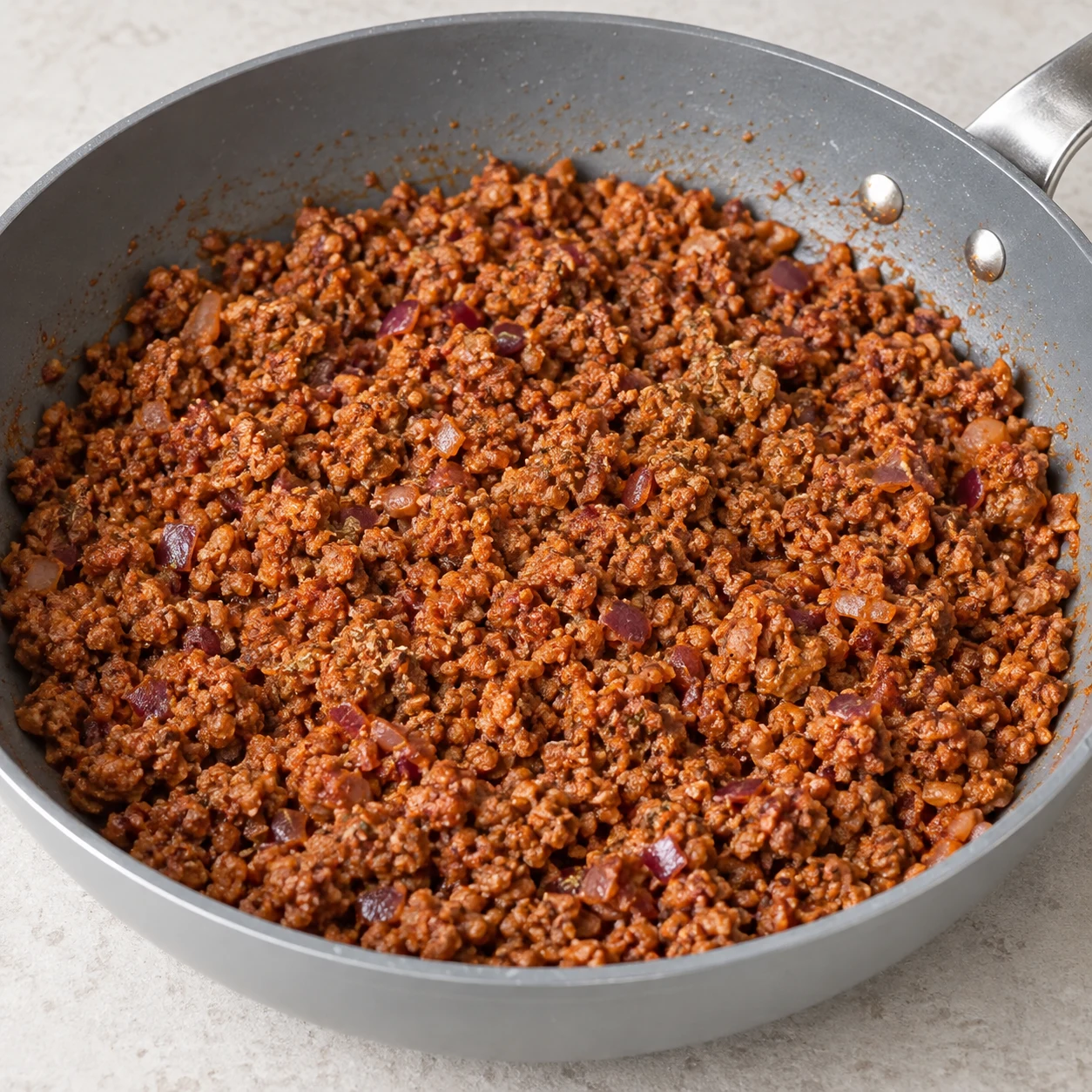 Incorpore les 60 g de concentré de tomate au haché assaisonné. Montrer seulement les ingrédients de cette phase ; mains anonymes, sauteuse grise et plan de travail pierre pâle.