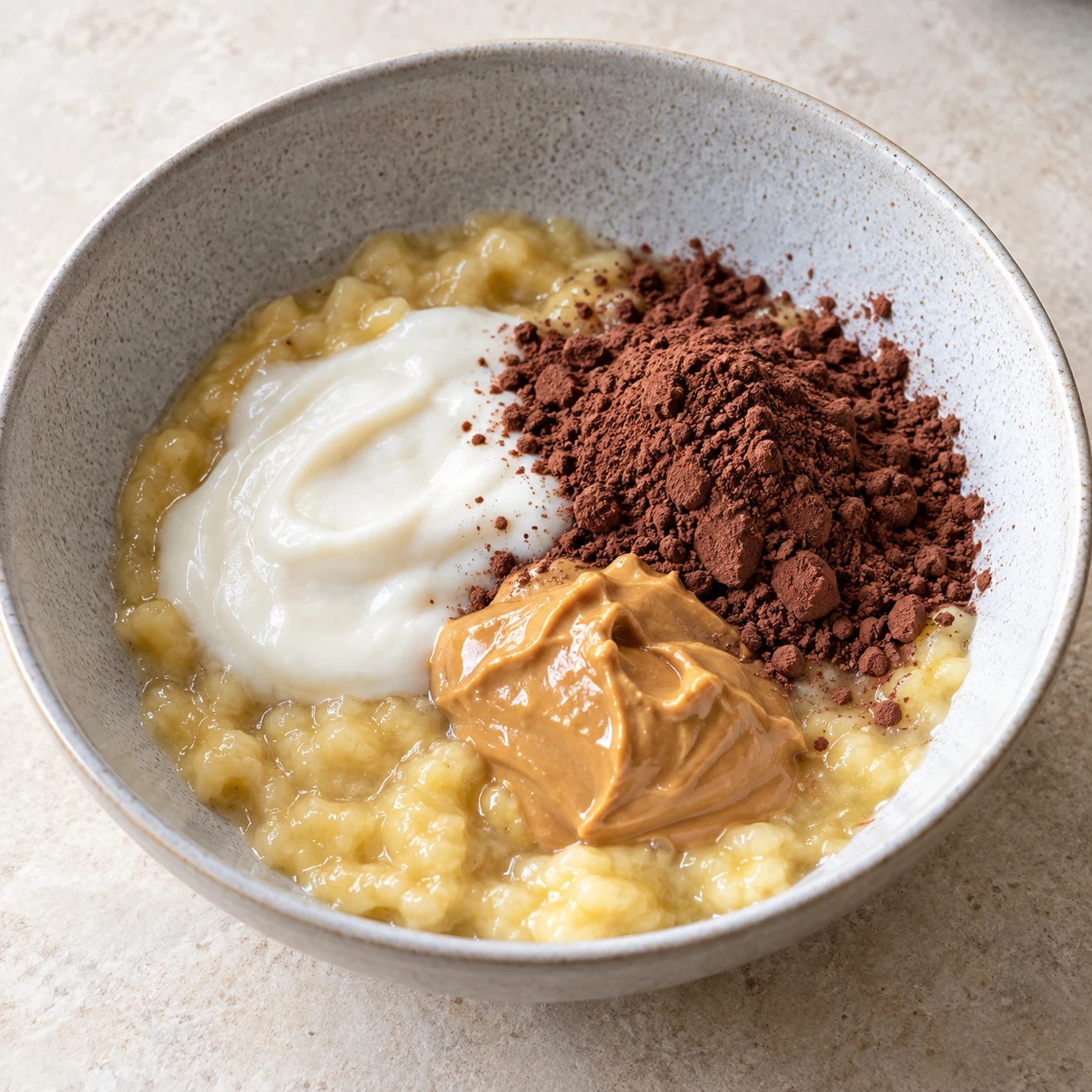 Bol contenant la banane écrasée, le yaourt blanc, le cacao brun et la purée de cacahuète ; ne pas ajouter de vanille visible.