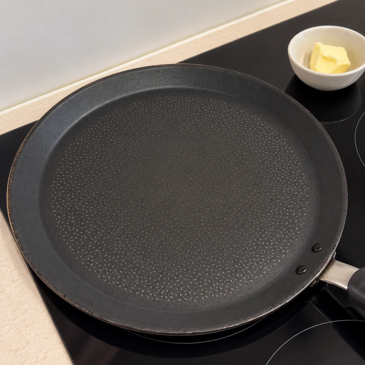 Poêle à crêpes noire vide sur une plaque ; petite noisette de margarine végétale dans une coupelle à côté, pas encore dans la poêle.