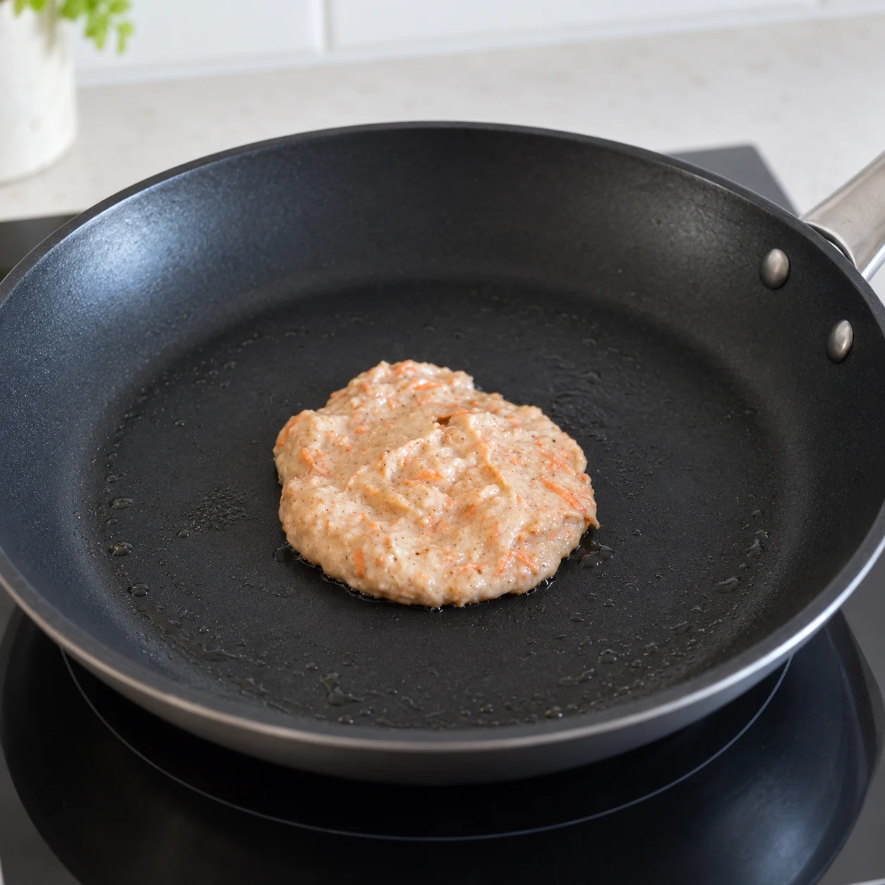 Petit pancake aux carottes dans poêle noire ; dessus encore humide et carotte râpée visible. Aucune spatule ni topping.