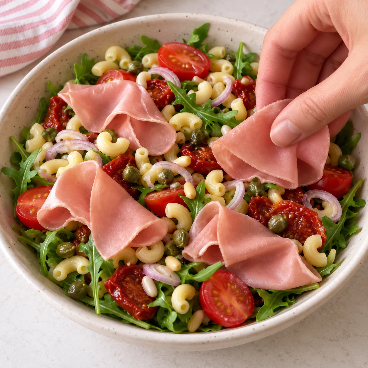 Une main anonyme dispose des fines tranches végétales roses uniformes de pois et fèves, sans gras animal, sur salade. Les pâtes éventuelles de cette adaptation sont des coquillettes courtes jaunes courbées et creuses, pas des radiateurs.