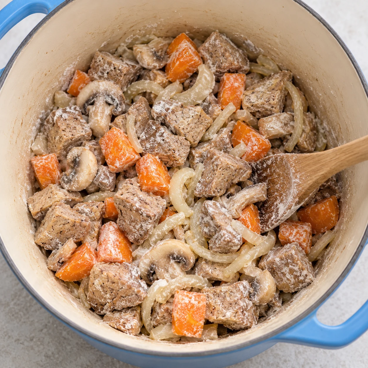 Vue de dessus d'une cocotte, farine blanche versée à la cuillère sur des morceaux de steak végétal, des tronçons de carotte, des champignons et des oignons.