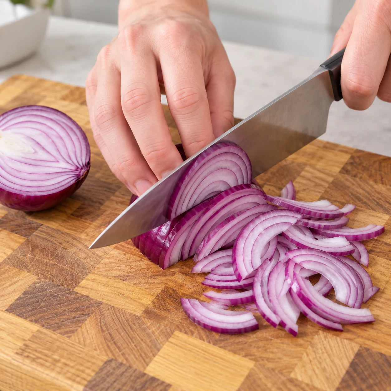 Oignon rouge coupé en deux sur une planche en bois, émincé en lamelles fines avec un couteau de chef, main adulte anonyme en griffe.