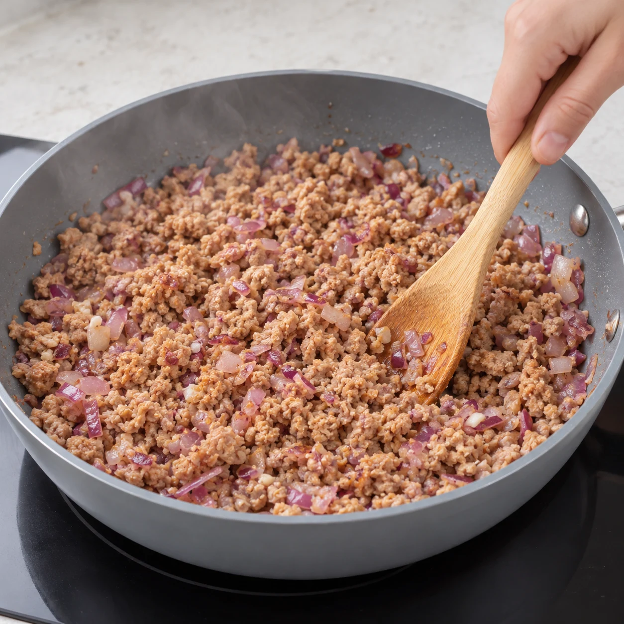 Ajoute les 300 g de haché végétal de soja cuisiné ; défais les amas et remue dans les oignons à feu modéré. Ce haché remplace le bœuf de la source. Montrer seulement les ingrédients de cette phase ; mains anonymes, sauteuse grise et plan de travail pierre pâle.