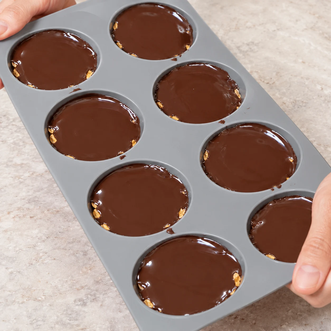 Mains anonymes tenant le moule légèrement incliné, chocolat liquide réparti jusqu’aux bords. La céréale croustillante est du corn flakes en fragments dorés et plats, jamais des grains de riz soufflé.