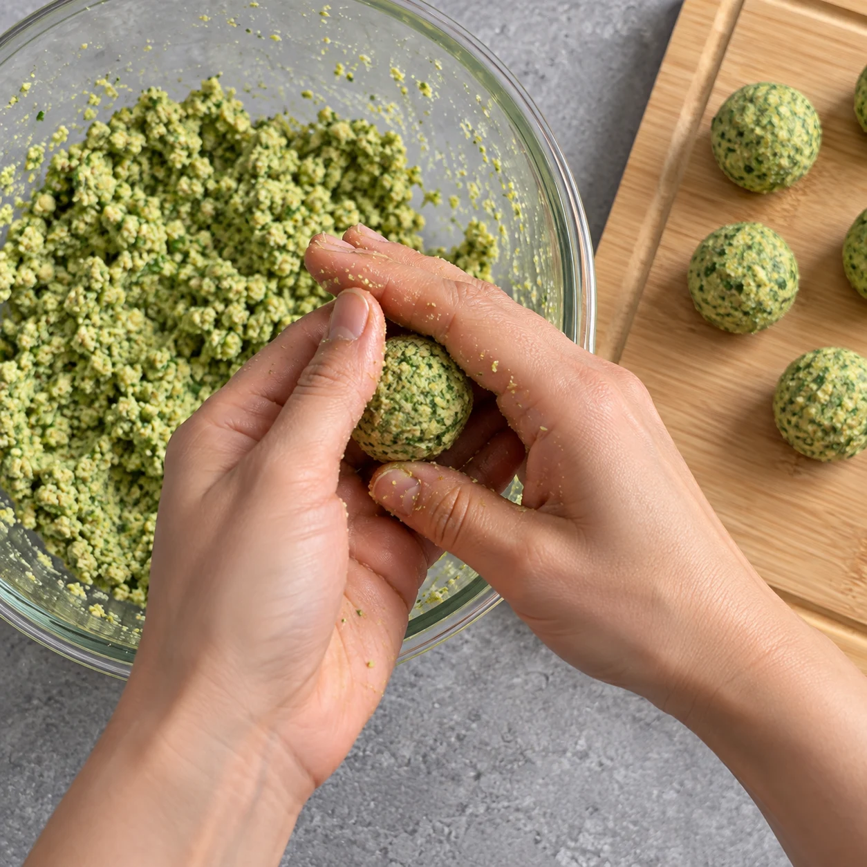Deux mains adultes anonymes façonnent une petite boule verte granuleuse au-dessus d’un saladier.