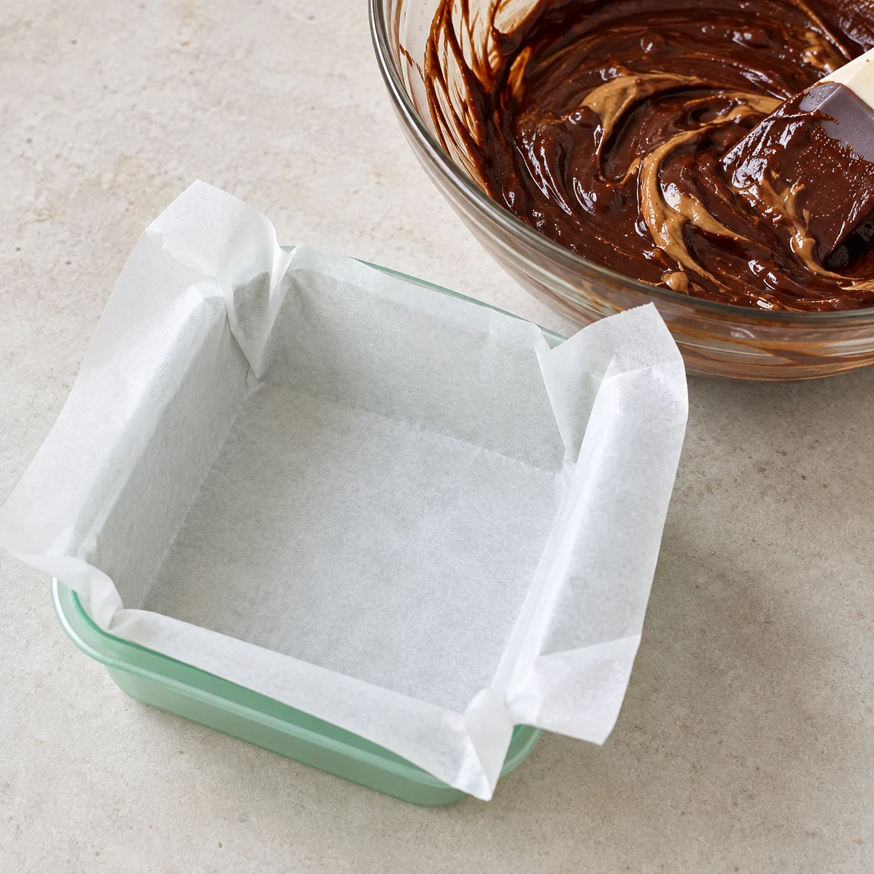 Boîte carrée chemisée de papier cuisson ; à côté, le bol de mélange chocolat-cacahuète attend d’être versé.