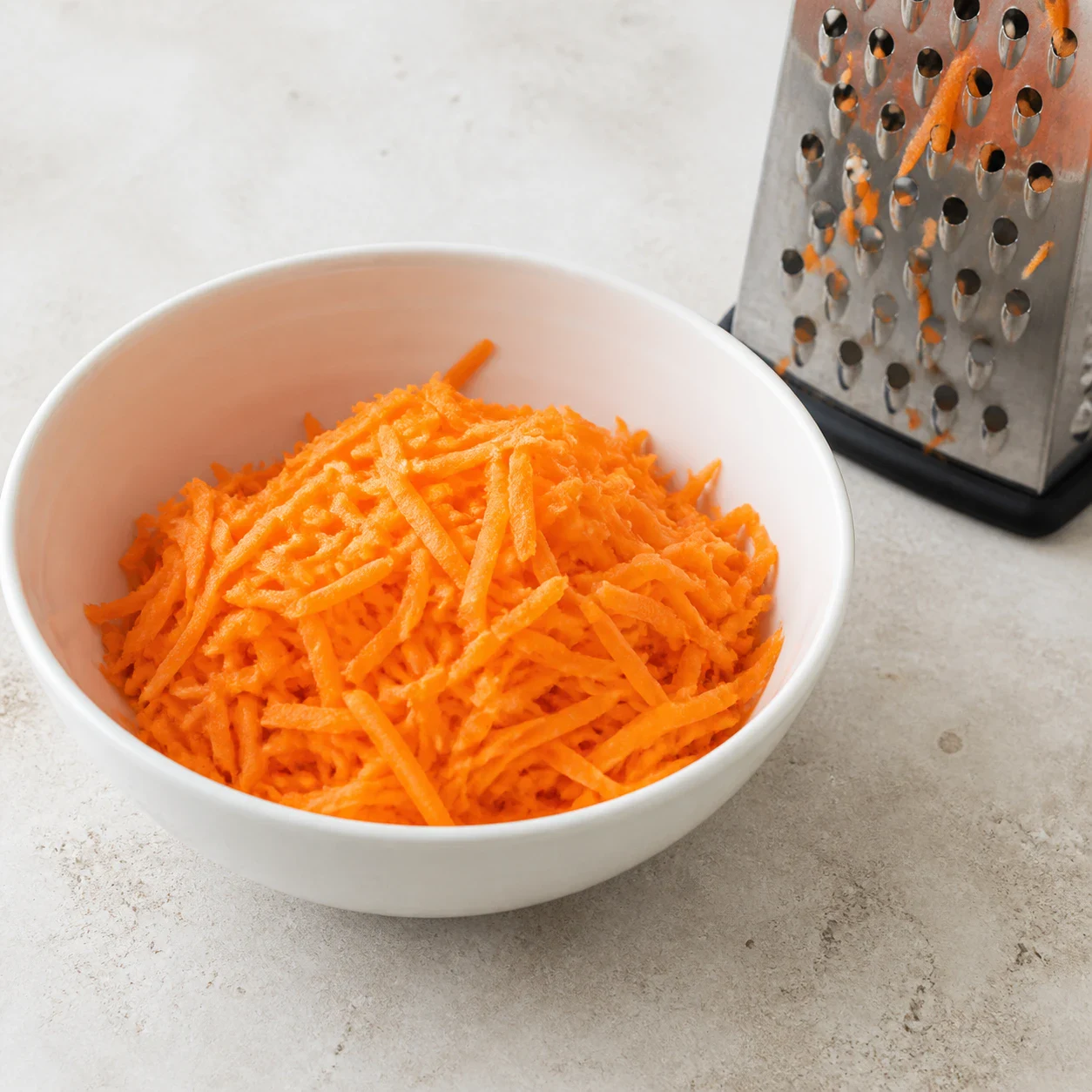 Saladier en céramique blanche contenant de la carotte râpée en filaments orange vif, râpe à gros trous posée à côté sur un plan de travail en pierre claire. Aucun autre ingrédient, aucune main, aucun texte.