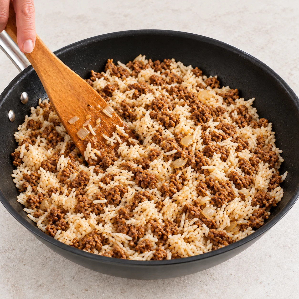 Incorporez le haché de soja chaud au riz et répartissez-le doucement à la spatule. Résultat visuel : Le haché de soja est réparti dans le riz. Montrer seulement les ingrédients de cette étape, sur plan de travail pierre pâle ; pas de viande, pas d’œuf, pas de tofu.